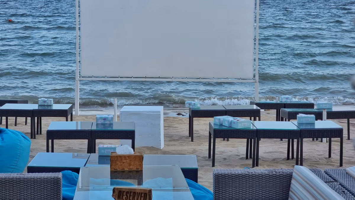 Pantalla de proyección montada en la playa frente a salas lounge y mesas bajas