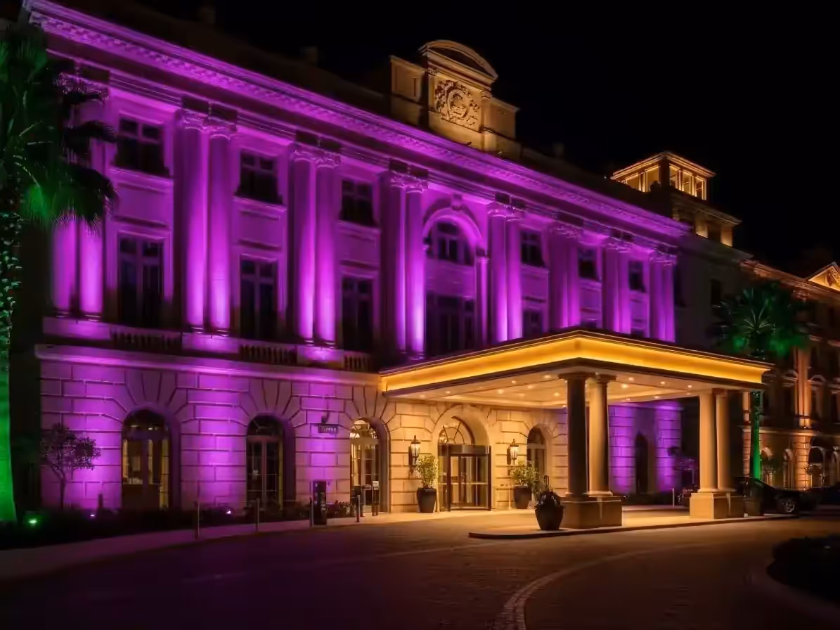 Fachada de hotel con columnas bañada en luz morada y acceso techado iluminado en blanco cálido, de noche