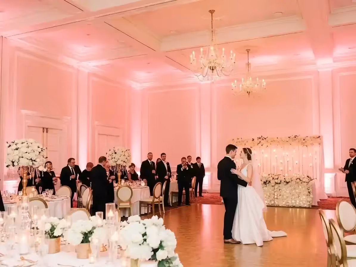Novios bailando en un salón con muros iluminados en rosa claro, candiles de cristal y centros de mesa con flores blancas