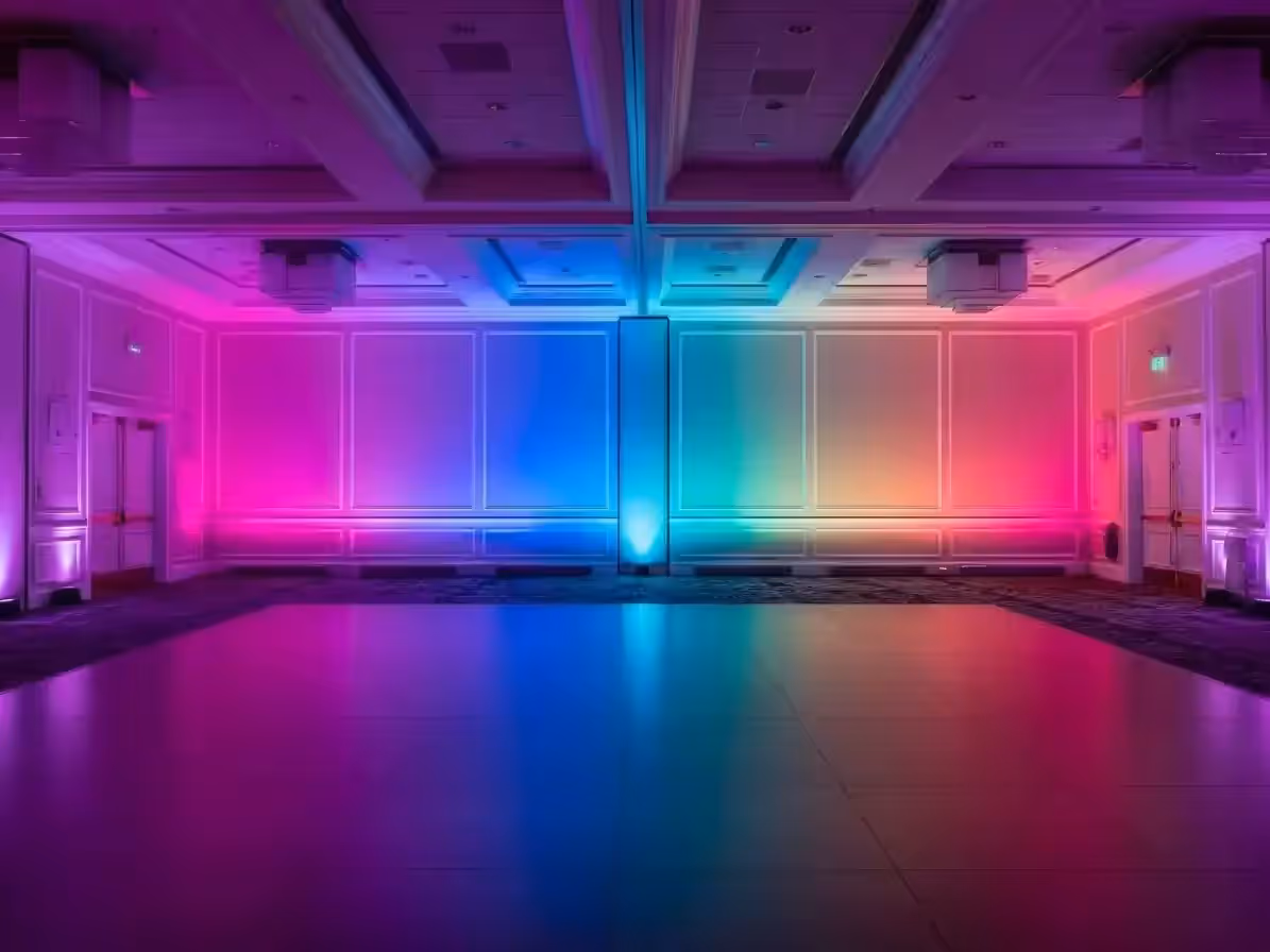 Salón vacío con muros iluminados desde el piso en un degradado de rosa, morado, azul y verde que se refleja en el piso de la pista