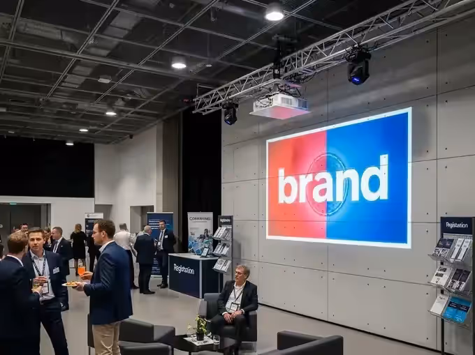 Logotipo a color proyectado sobre un muro gris en un espacio corporativo con estructura de truss y asistentes de traje