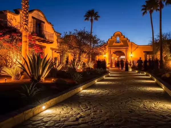 Camino de entrada de una hacienda mexicana con agaves y palmeras iluminados desde el piso