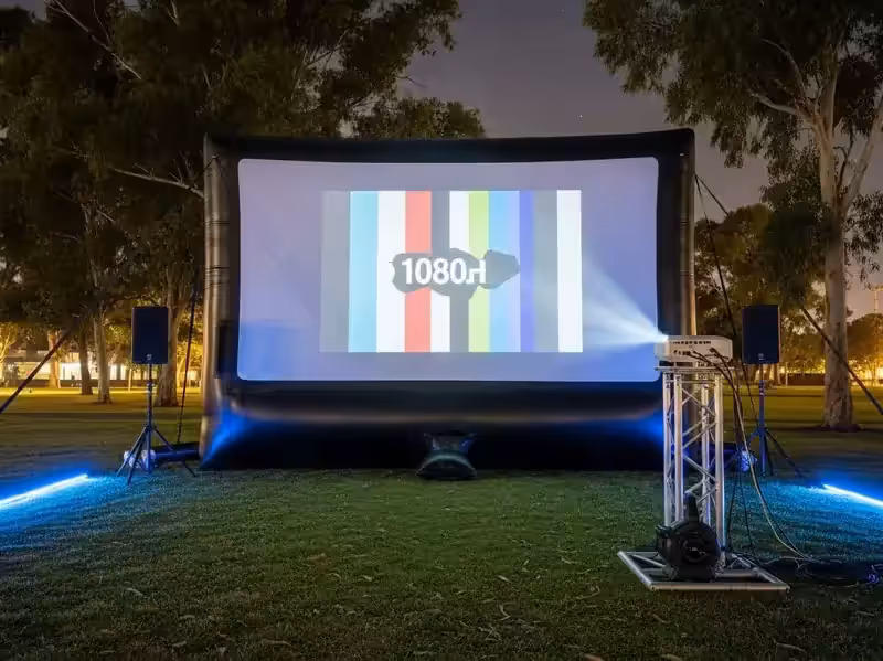 Pantalla inflable de noche en un parque con patrón de prueba en pantalla, proyector sobre torre de truss y dos bocinas en tripié